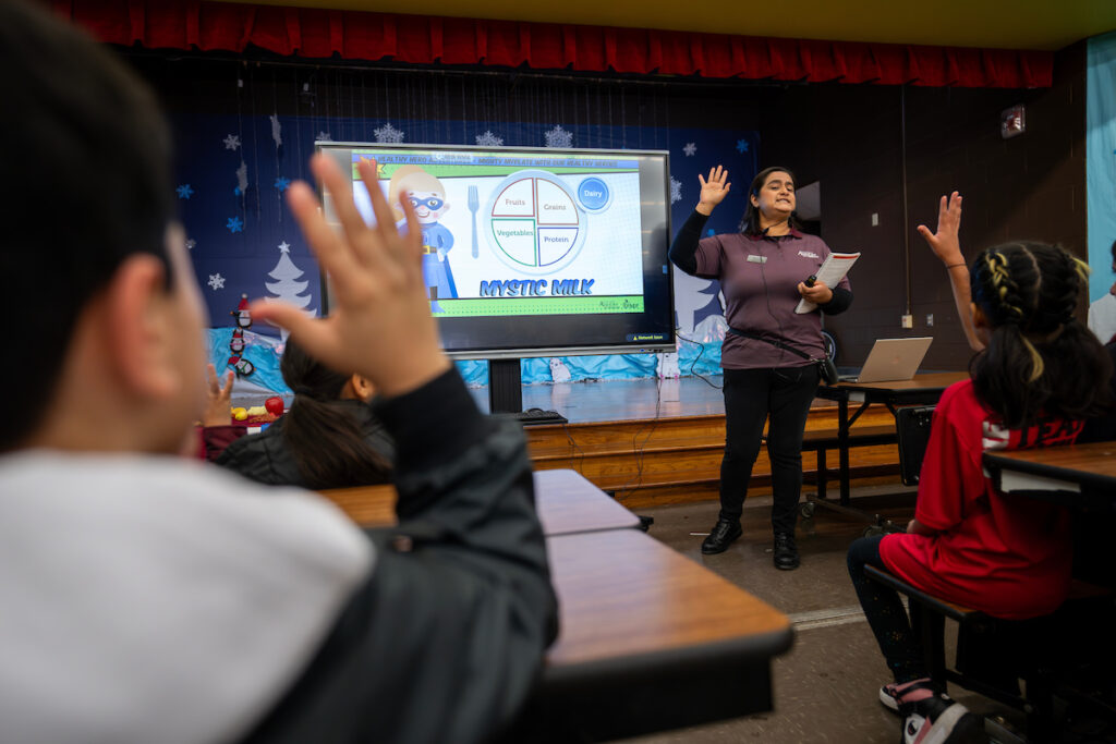 A woman in front of a projector screen wearing an AgriLife Extension shirt raising her hand along with a room full of 4th grade students