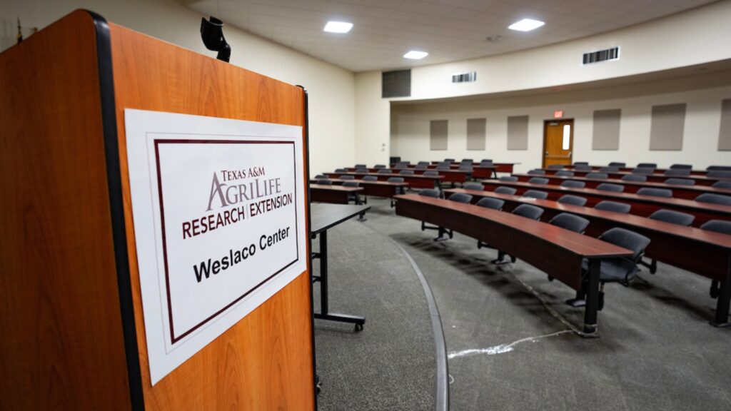 Close up of podium showing Texas A&M AgriLife Research and Extension Center at Weslaco logo. Rows of seats and long tables out of focus in the background